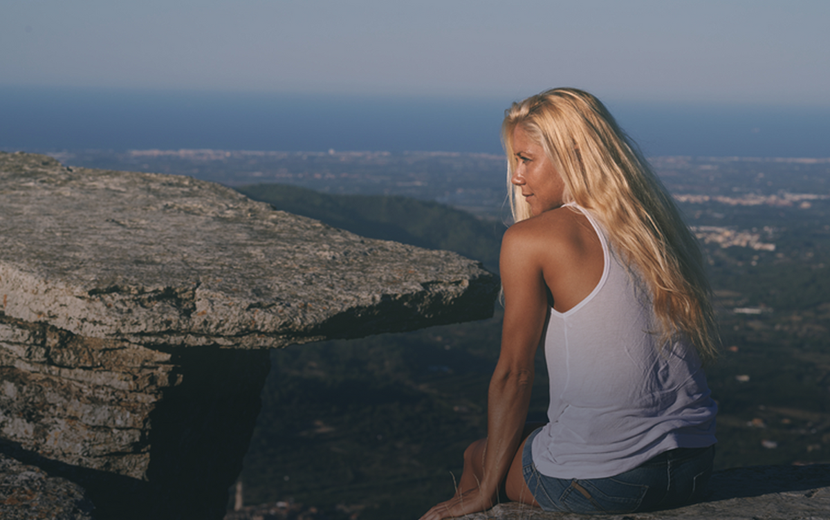 A woman with long blonde hair sits on a rocky cliffside overlooking a vast landscape and the sea under a clear blue sky, wearing a white tank top and denim shorts.